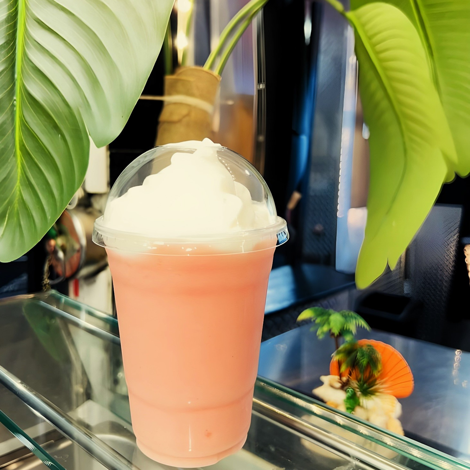 A pink Caribbean smoothie with whipped cream in a plastic cup, surrounded by tropical leaves.