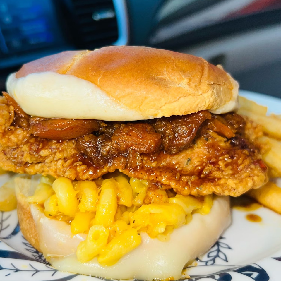 A Caribbean fried chicken sandwich with mac and cheese, caramelized onions, and fries on a plate.