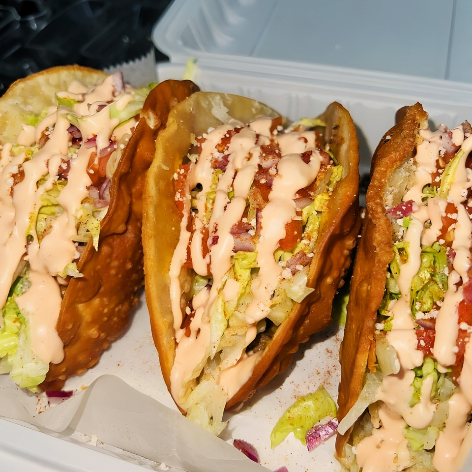 Three Caribbean tacos with fried shells, lettuce, tomatoes, and a creamy pink sauce in a takeout container.